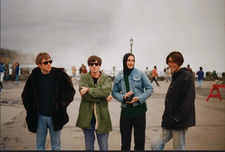 Four young men in coats standing on a grey seafront, one holding a camera