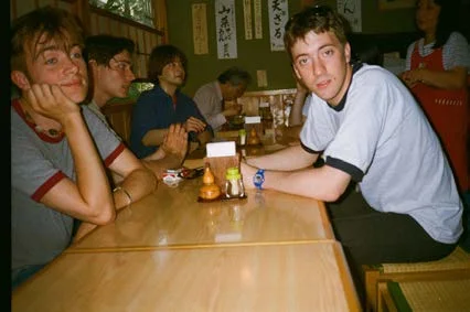 Young men sitting at a long wooden restaurant table with Japanese signs on the wall behind