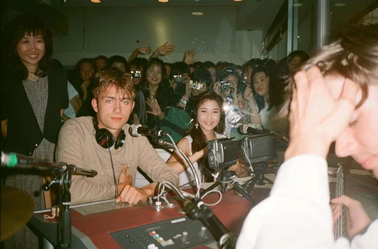 A young man in headphones at a microphone desk, with a crowd of people holding cameras behind a glass screen