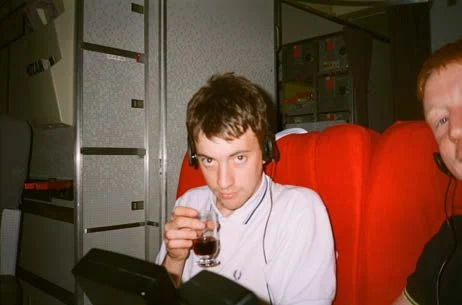 A young man in headphones holding a small glass, seated in a red seat