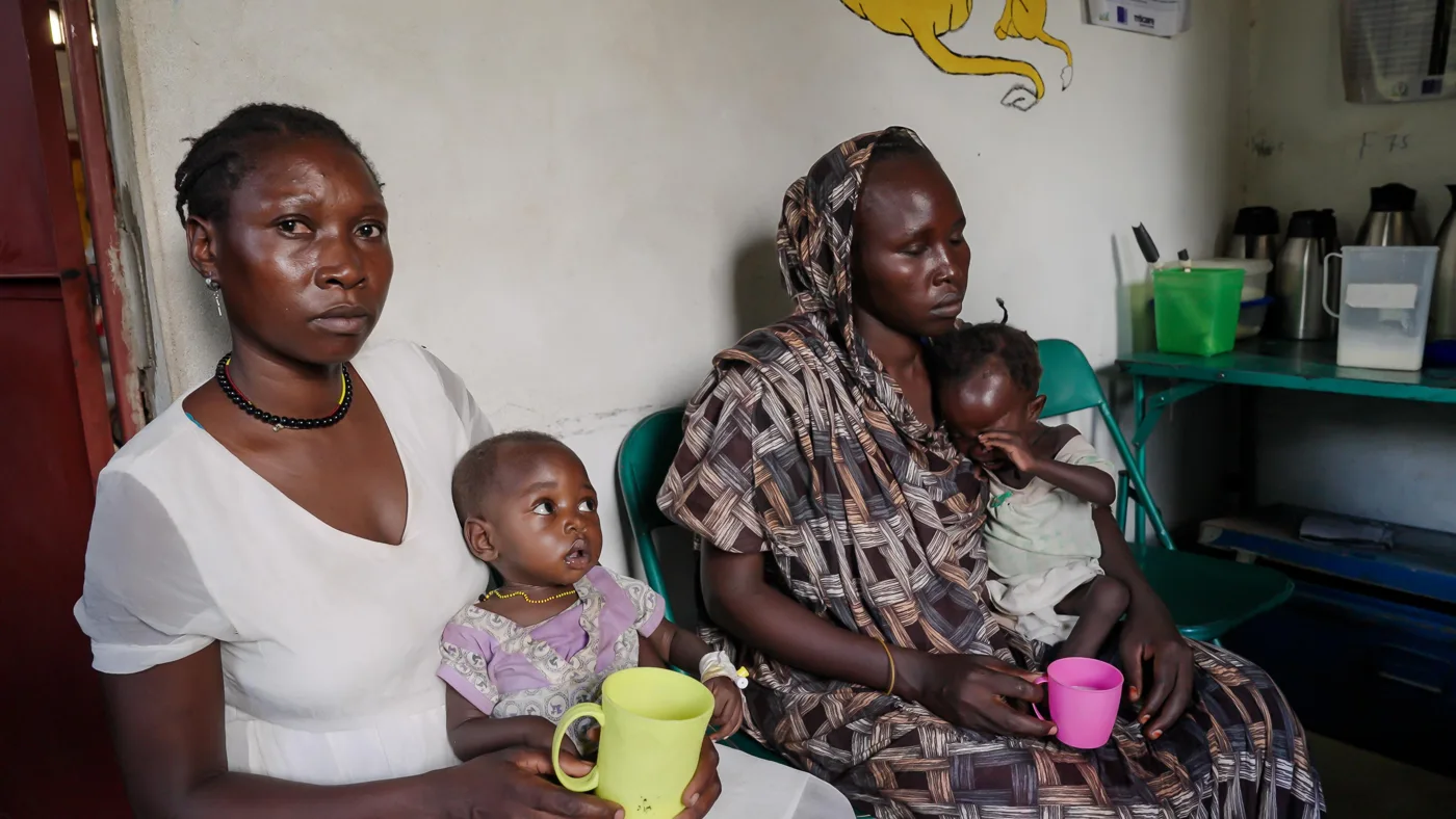 Two women seated against a white wall, each holding a young child, one child holding a green cup and the other a pink cup
