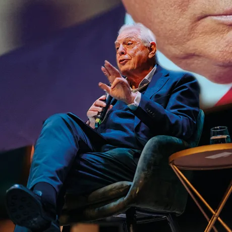 John Simpson speaking into a microphone from an armchair on stage