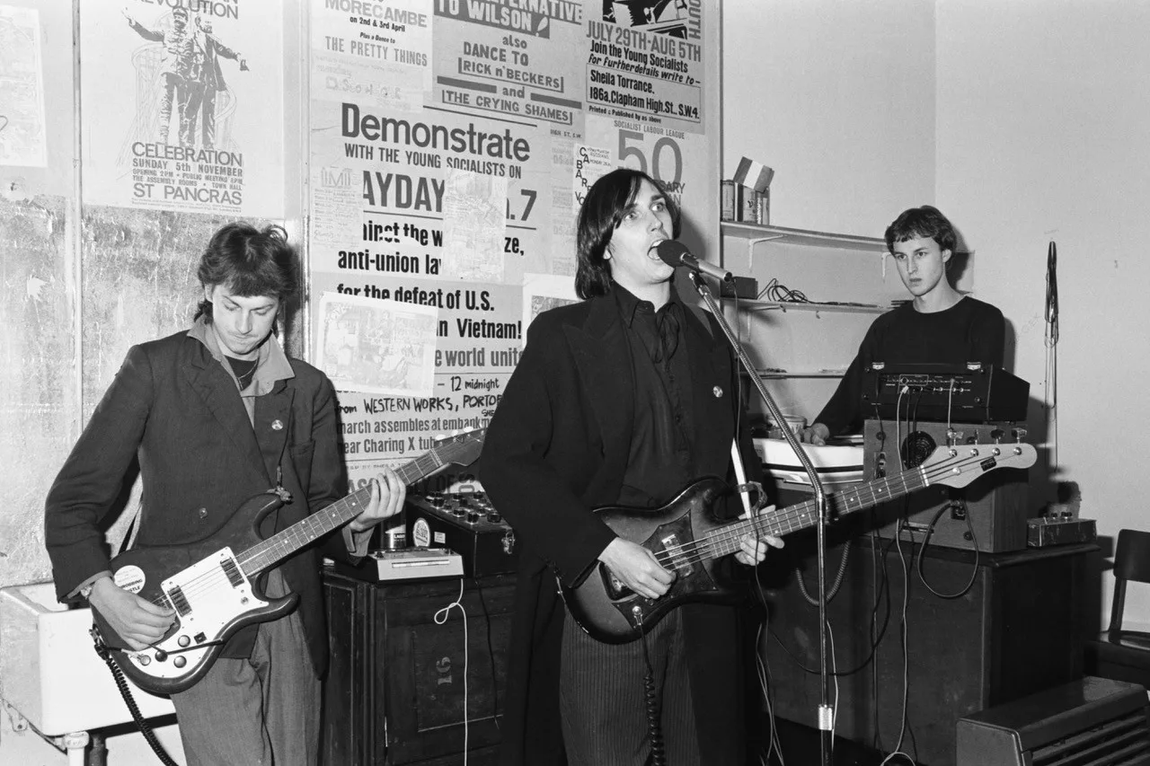 Black and white photograph of three young men performing with guitars and a synthesiser in front of a wall of political posters