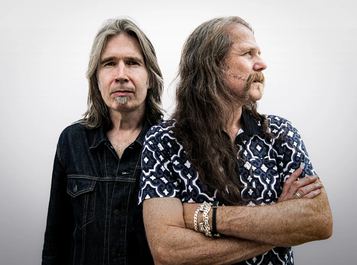 Two long-haired men photographed against a pale grey background, one in a denim jacket, one with arms folded